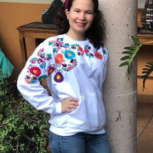 Puede incluir: Una sudadera blanca con bordado floral de colores alrededor del cuello. La sudadera tiene un bolsillo canguro y mangas largas. La persona que lleva la sudadera est&aacute; de pie junto a un pilar.