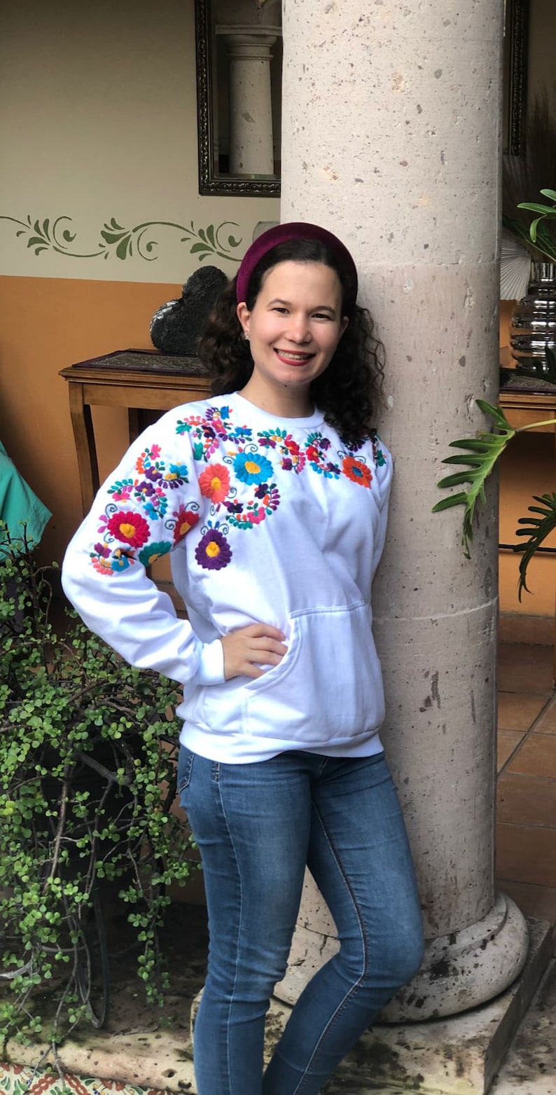 Puede incluir: Una sudadera blanca con bordado floral de colores alrededor del cuello. La sudadera tiene un bolsillo canguro y mangas largas. La persona que lleva la sudadera est&aacute; de pie junto a un pilar.