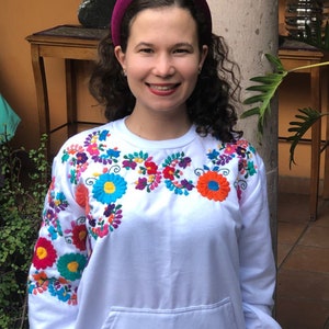 Puede incluir: Sudadera blanca con flores bordadas de colores en el pecho y las mangas. La sudadera tiene un bolsillo delantero y un corte holgado.