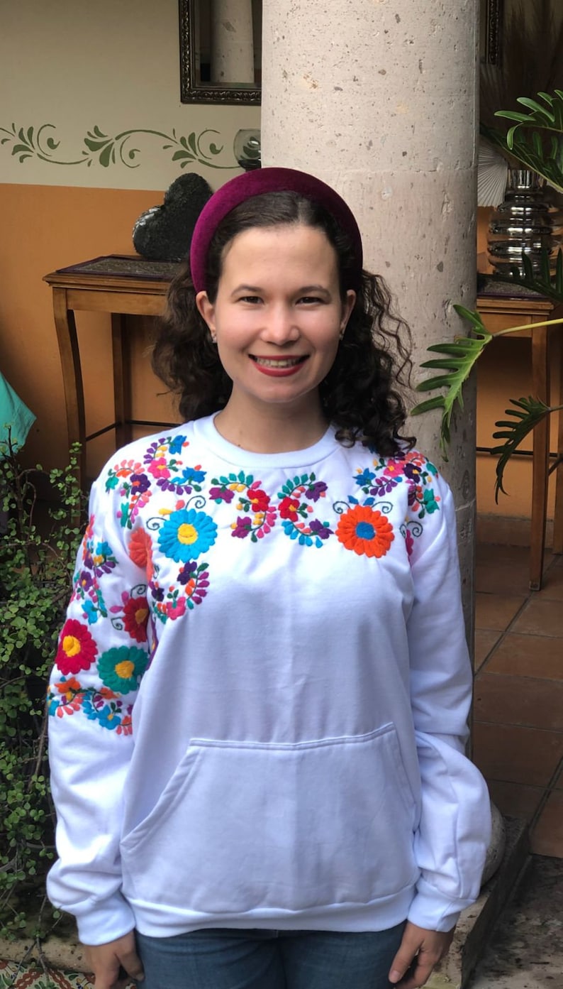Puede incluir: Sudadera blanca con flores bordadas de colores en el pecho y las mangas. La sudadera tiene un bolsillo delantero y un corte holgado.