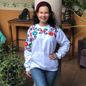 Puede incluir: Una sudadera blanca con flores bordadas de colores alrededor del cuello. La sudadera tiene un bolsillo canguro y mangas largas. La modelo lleva jeans azules y una diadema color burdeos.