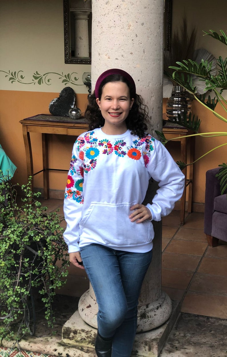 Puede incluir: Una sudadera blanca con flores bordadas de colores alrededor del cuello. La sudadera tiene un bolsillo canguro y mangas largas. La modelo lleva jeans azules y una diadema color burdeos.