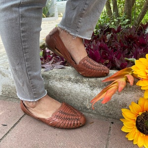 May include: A pair of brown leather woven sandals with a criss-cross pattern. The sandals are worn by a person wearing grey jeans.