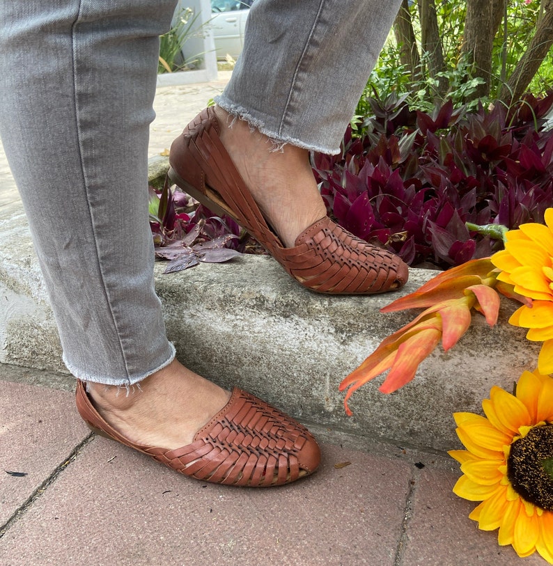 May include: A pair of brown leather woven sandals with a criss-cross pattern. The sandals are worn by a person wearing grey jeans.