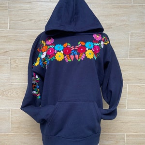 May include: A navy blue hooded sweatshirt with colorful floral embroidery on the front and sleeve. The embroidery features a variety of flowers in shades of pink, yellow, red, blue, and green.