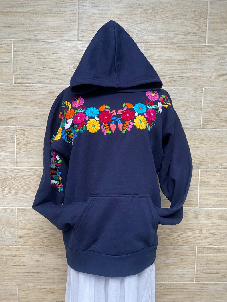 May include: A navy blue hooded sweatshirt with colorful floral embroidery on the front and sleeve. The embroidery features a variety of flowers in shades of pink, yellow, red, blue, and green.