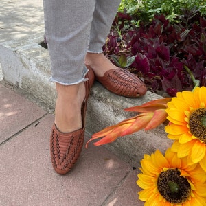 May include: A pair of brown leather woven sandals with a closed toe and an open heel. The sandals are worn by a person standing on a concrete surface with sunflowers in the background.