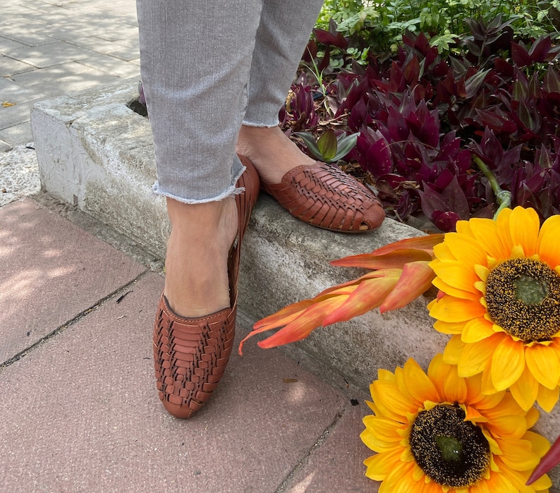 May include: A pair of brown leather woven sandals with a closed toe and an open heel. The sandals are worn by a person standing on a concrete surface with sunflowers in the background.