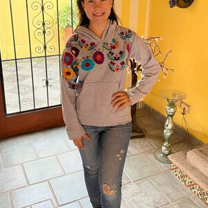 May include: A gray hoodie with colorful embroidered flowers on the front. The hoodie has a hood and a kangaroo pocket. The person is wearing blue jeans and brown shoes.