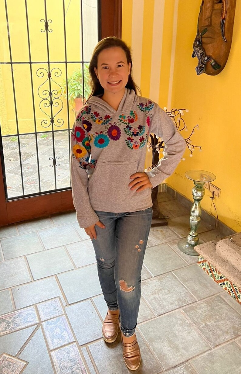 May include: A gray hoodie with colorful embroidered flowers on the front. The hoodie has a hood and a kangaroo pocket. The person is wearing blue jeans and brown shoes.