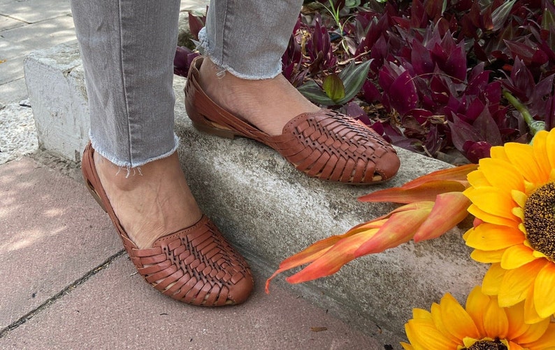 May include: A pair of brown leather woven sandals with a flat sole. The sandals are worn by a person standing on a concrete curb with a sunflower in the background.