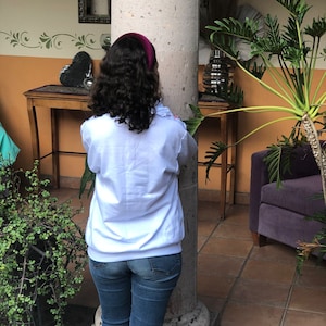 Puede incluir: Una mujer con el pelo largo y rizado casta&ntilde;o lleva una sudadera blanca y unos vaqueros azules. Est&aacute; de pie frente a un pilar y mirando hacia la derecha.
