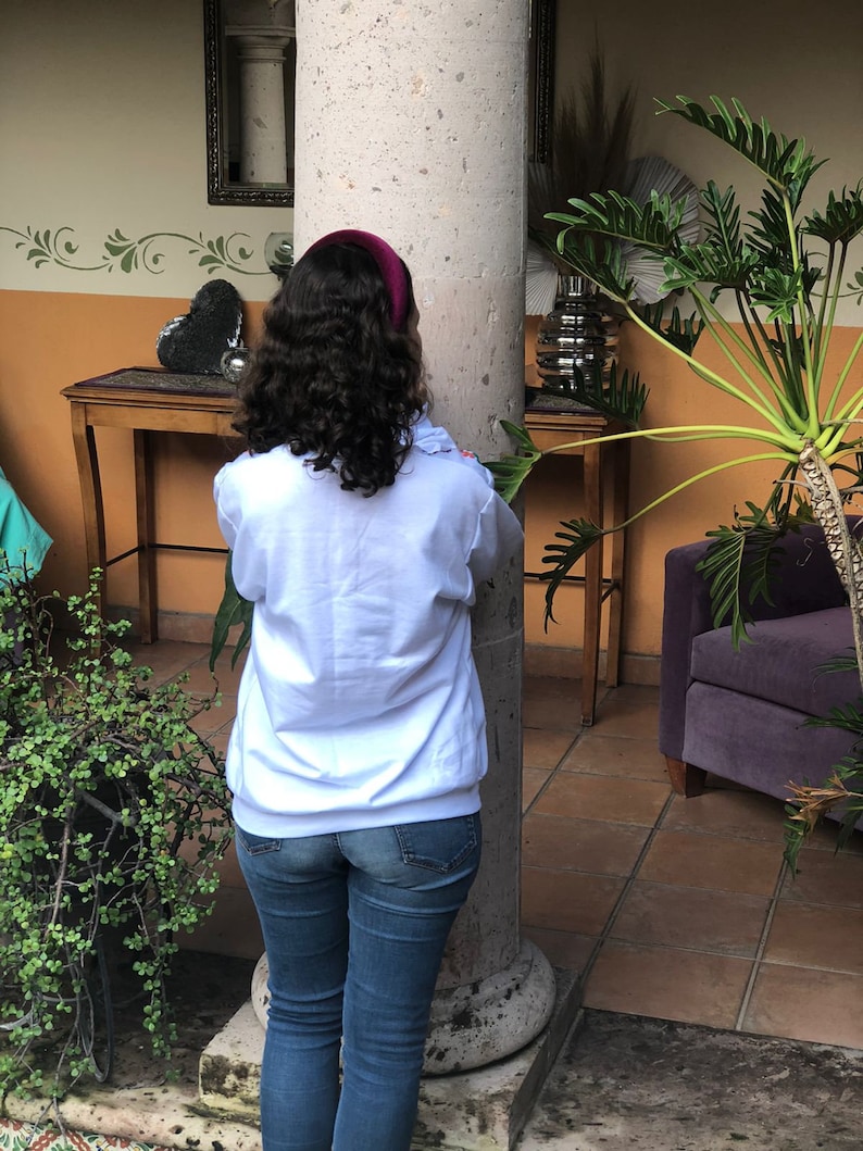 Puede incluir: Una mujer con el pelo largo y rizado casta&ntilde;o lleva una sudadera blanca y unos vaqueros azules. Est&aacute; de pie frente a un pilar y mirando hacia la derecha.