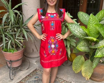 May include: A woman wearing a red embroidered dress with colorful floral patterns. She is standing in a courtyard with green plants and a stone floor.