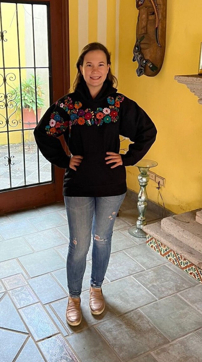 May include: A woman wearing a black hoodie with colorful embroidered flowers on the chest, blue jeans, and brown loafers. She is standing in a room with a tiled floor and a yellow wall.