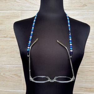 May include: A beaded glasses chain with blue, white, and light blue beads. The chain is hanging from a black mannequin's neck and is holding up a pair of clear glasses.