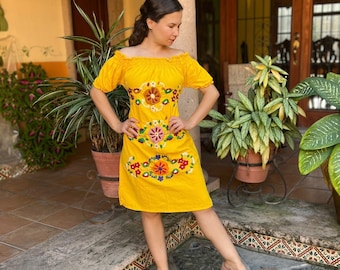 May include: A yellow off-the-shoulder dress with colorful floral embroidery. The dress features short sleeves and a knee-length hemline. The embroidery includes orange, pink, blue, and green flowers. The dress is made of a lightweight fabric.