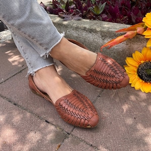 May include: A pair of brown leather sandals with a woven design. The sandals are worn by a person with bare feet. The sandals are on a brick walkway.