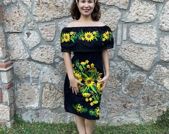 May include: A black off-the-shoulder dress with embroidered sunflowers and green leaves. The dress has a ruffled neckline and a fitted silhouette. The model is wearing a white headband.