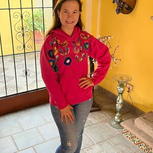 May include: A pink hoodie with colorful embroidered flowers on the front. The hoodie has a hood and a kangaroo pocket. The person wearing the hoodie is standing in a room with tiled floors.