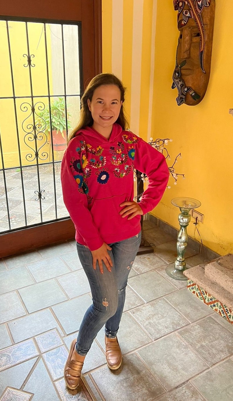 May include: A pink hoodie with colorful embroidered flowers on the front. The hoodie has a hood and a kangaroo pocket. The person wearing the hoodie is standing in a room with tiled floors.