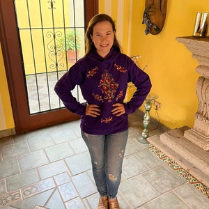 May include: A purple hoodie with colorful embroidery on the front. The hoodie has a drawstring hood and long sleeves. The person is wearing blue jeans and brown shoes.