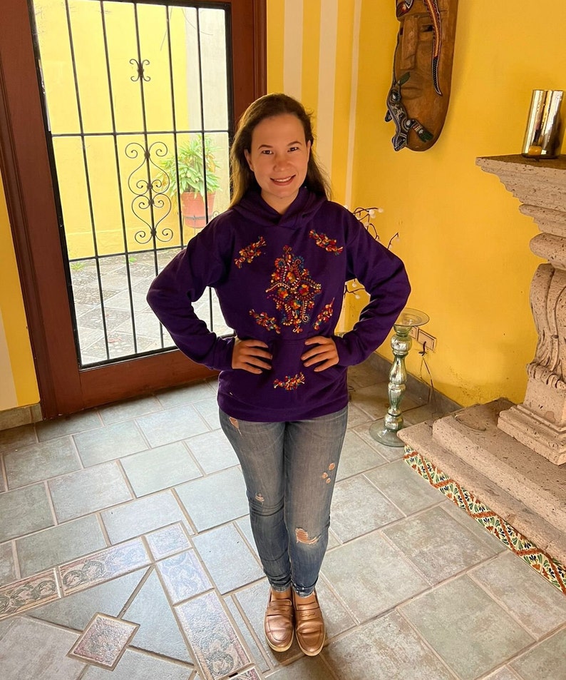 May include: A purple hoodie with colorful embroidery on the front. The hoodie has a drawstring hood and long sleeves. The person is wearing blue jeans and brown shoes.