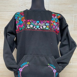 Puede incluir: Sudadera negra con un gran dise&ntilde;o floral bordado en el pecho y un dise&ntilde;o floral m&aacute;s peque&ntilde;o en el bolsillo. La sudadera tiene mangas largas y cuello redondo.