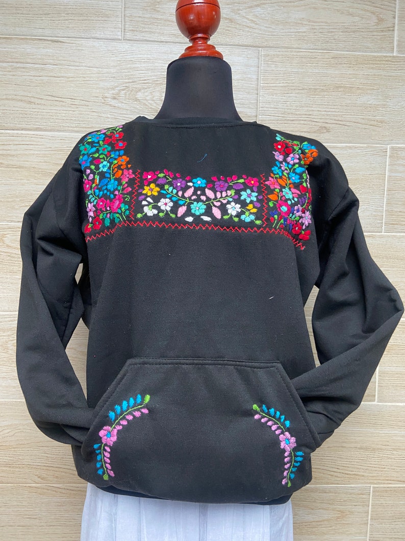 Puede incluir: Sudadera negra con un gran dise&ntilde;o floral bordado en el pecho y un dise&ntilde;o floral m&aacute;s peque&ntilde;o en el bolsillo. La sudadera tiene mangas largas y cuello redondo.