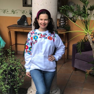 Puede incluir: Una sudadera blanca con flores bordadas de colores alrededor del cuello. La sudadera tiene un bolsillo en la parte delantera y la lleva puesta una persona con una diadema color burdeos.