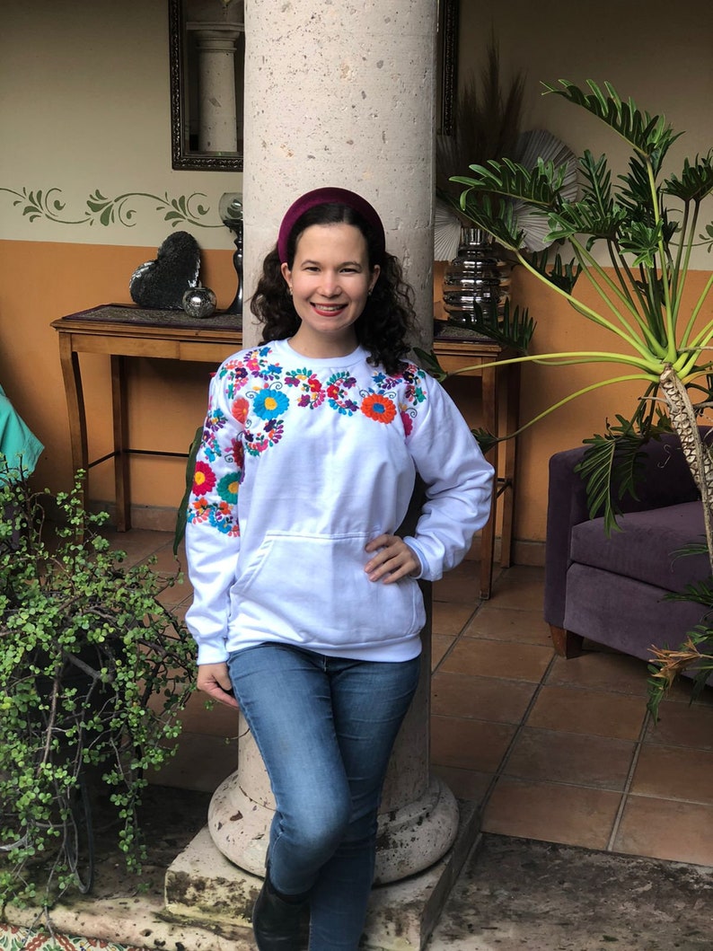 Puede incluir: Una sudadera blanca con flores bordadas de colores alrededor del cuello. La sudadera tiene un bolsillo en la parte delantera y la lleva puesta una persona con una diadema color burdeos.