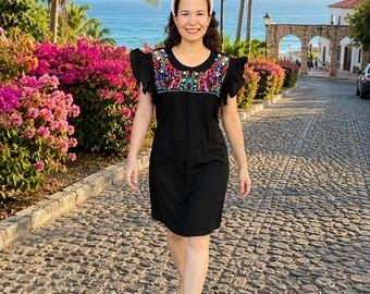 May include: A black dress with colorful floral embroidery around the neckline and on the sleeves. The dress has a round neckline and short sleeves. The person is wearing a pink headband and beige sandals. The background includes palm trees, flowers, and a cobblestone street.