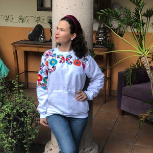 Puede incluir: Una sudadera blanca con flores bordadas de colores en la parte delantera y las mangas. La sudadera tiene un bolsillo canguro y la lleva puesta una persona con una diadema color burdeos.