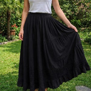 May include: A white sleeveless vest top with a scoop neckline paired with a long, black, tiered maxi skirt. The skirt features a gathered elastic waistband, multiple horizontal stitched panels, and a ruffled hemline that reaches the ankles.