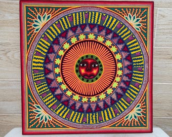 Puede incluir: Una obra de arte circular colorida con una cara de sol estilizada en el centro. El sol tiene una cara roja con ojos negros y una boca. La obra de arte está rodeada de círculos concéntricos de amarillo, verde, azul y rojo.