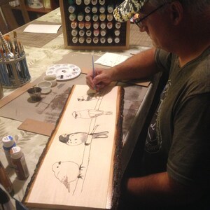 May include: A person is painting a wood slice with a bird design. The artwork features several birds perched on a line, rendered in black ink. The wood slice has a natural bark edge. Paint supplies and a paint organizer are visible.