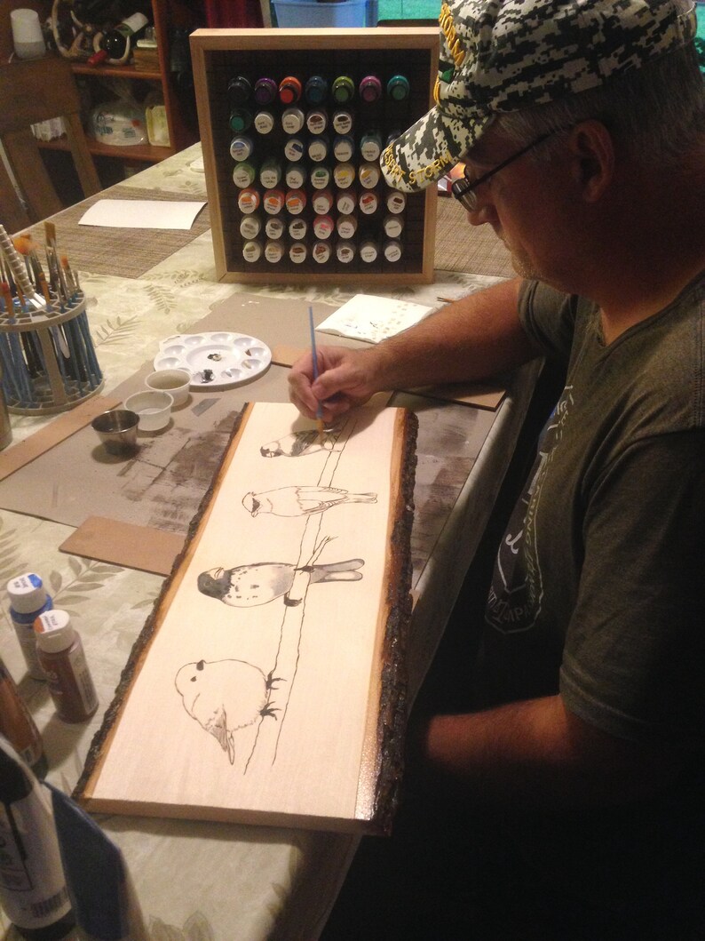 May include: A person is painting a wood slice with a bird design. The artwork features several birds perched on a line, rendered in black ink. The wood slice has a natural bark edge. Paint supplies and a paint organizer are visible.