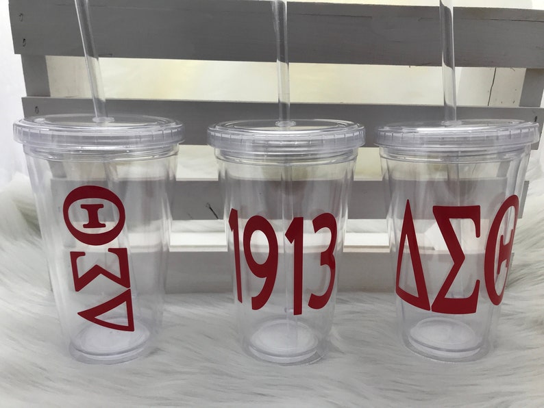 May include: Three clear plastic tumblers with lids and straws. Each tumbler has red lettering: one with Greek letters, one with the number 1913, and one with Greek letters. The tumblers are on a white surface.