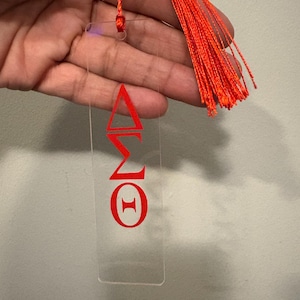 May include: Clear acrylic bookmark with red Greek letters and a bright orange tassel. The bookmark features the Greek letters Delta, Sigma, and Theta. The tassel is attached to the top with a matching orange cord.