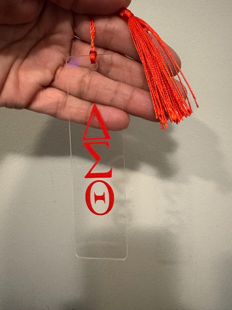 May include: Clear acrylic bookmark with red Greek letters and a bright orange tassel. The bookmark features the Greek letters Delta, Sigma, and Theta. The tassel is attached to the top with a matching orange cord.