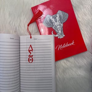 May include: A red notebook with the word "Notebook" and a detailed elephant illustration. A clear bookmark with red Greek letters is inside the notebook. A red tassel is attached to the bookmark.