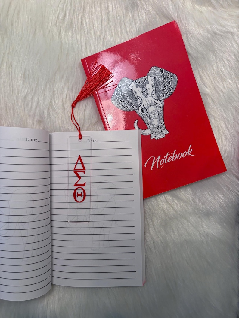 May include: A red notebook with the word "Notebook" and a detailed elephant illustration. A clear bookmark with red Greek letters is inside the notebook. A red tassel is attached to the bookmark.