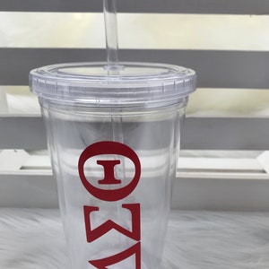 May include: A clear plastic tumbler with a lid and straw. The tumbler features red Greek letters. The design is simple and modern, perfect for cold beverages. The tumbler is made of durable plastic and is easy to clean.