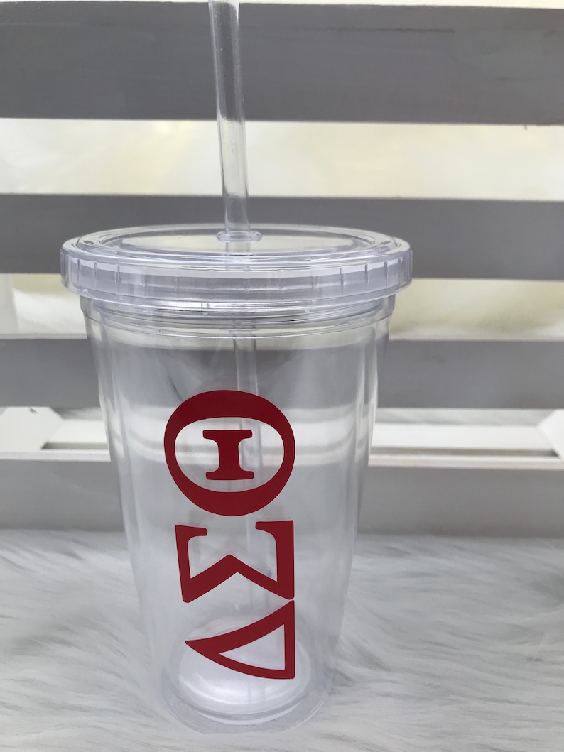 May include: A clear plastic tumbler with a lid and straw. The tumbler features red Greek letters. The design is simple and modern, perfect for cold beverages. The tumbler is made of durable plastic and is easy to clean.