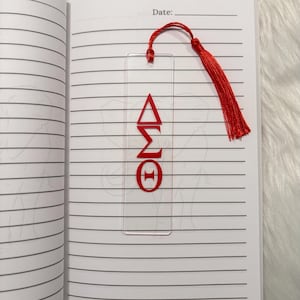 May include: Clear acrylic bookmark with red Greek letters &Delta;&Sigma;&Theta; and a red tassel. The bookmark is placed in an open notebook with lined pages. The bookmark is a decorative accessory for books and journals.