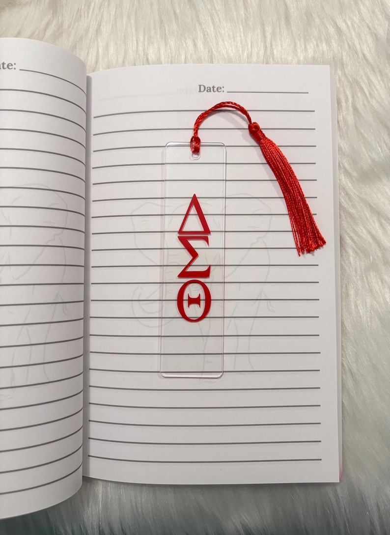 May include: Clear acrylic bookmark with red Greek letters &Delta;&Sigma;&Theta; and a red tassel. The bookmark is placed in an open notebook with lined pages. The bookmark is a decorative accessory for books and journals.