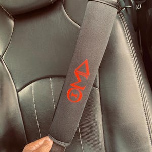 May include: Black car seatbelt cover with a red logo that says "ONE" with a triangle above the "O".