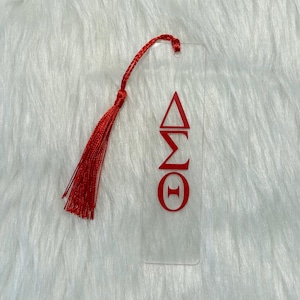 May include: Clear acrylic bookmark with red Greek letters and a red tassel. The bookmark features the Greek letters Delta, Sigma, and Theta. The tassel is attached at the top with a small loop. The bookmark is rectangular and has a smooth surface.