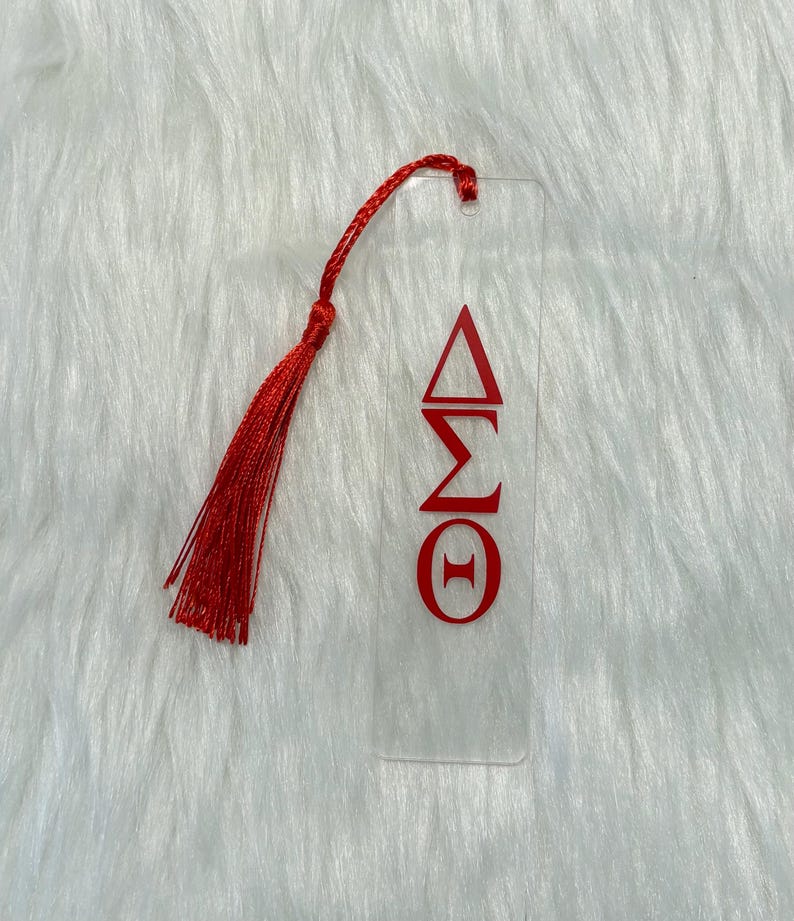May include: Clear acrylic bookmark with red Greek letters and a red tassel. The bookmark features the Greek letters Delta, Sigma, and Theta. The tassel is attached at the top with a small loop. The bookmark is rectangular and has a smooth surface.