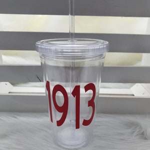 May include: Clear plastic tumbler with a lid and straw. The tumbler features the number "1913" in a bold, red font. The cup is designed for cold beverages and is shown on a white, fluffy surface.